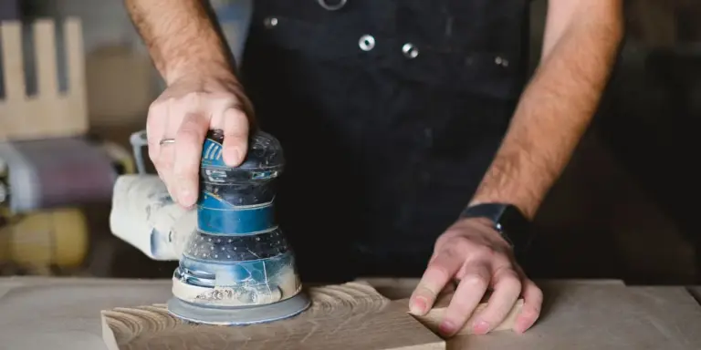Ein Exzenterschleifer auf einer Holzfläche, die zweite Hand hält das Werkstück