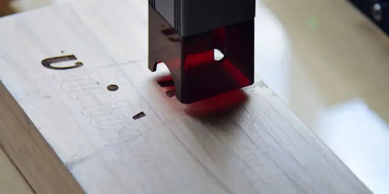 Ein Laserkopf steht über einem hellen Holzbrett, in das bereits Schlitze geschnitten sind