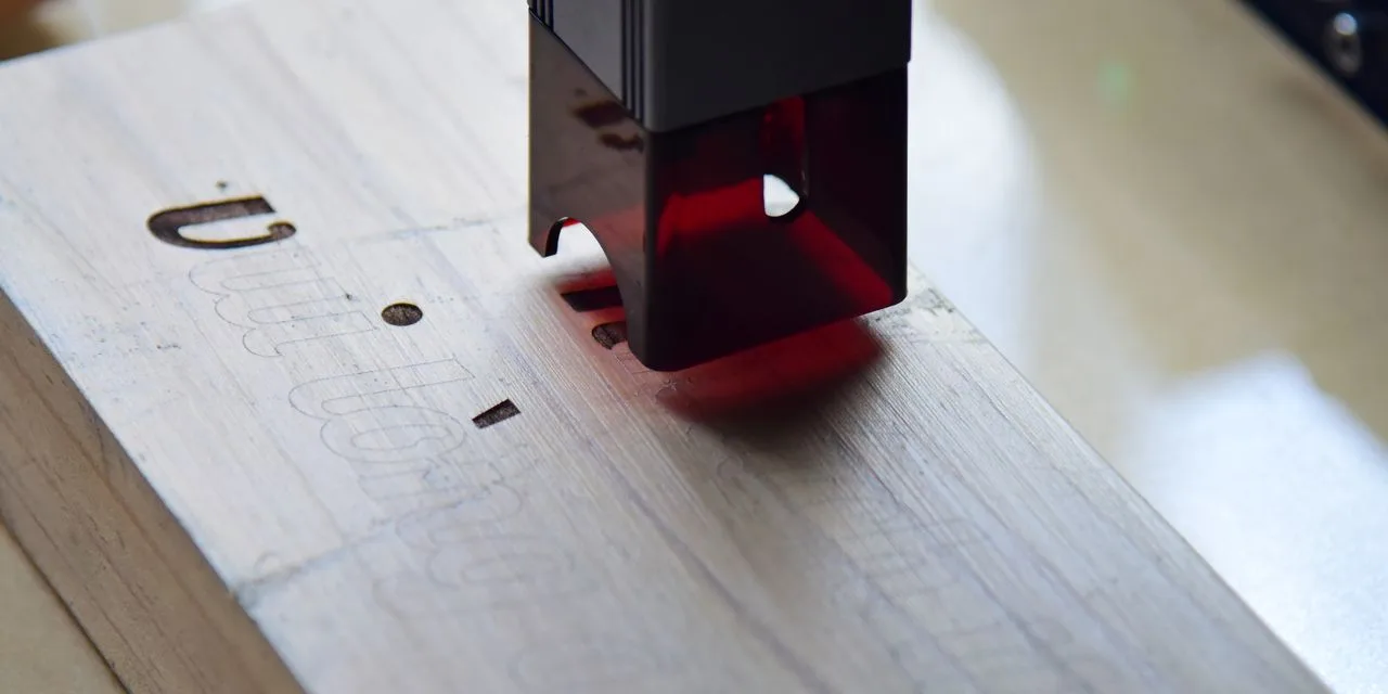 Ein Laserkopf steht über einem hellen Holzbrett, in das bereits Schlitze geschnitten sind